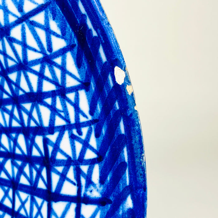 Antique Manises majolica charger with a large circular form, shallow well, and broad decorated rim. Saturated cobalt brushwork creates a woven lattice of intersecting lines, surrounding a white central reserve with simplified foliage and geometric marks. A visible signature and natural edge wear distinguish this Spanish ceramic wall plate.