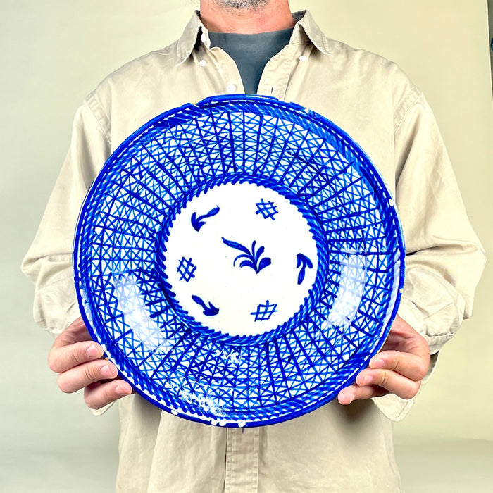 Hand painted blue charger from Manises, Spain, formed in glazed earthenware with a wide sloping rim and low profile. Cobalt lines overlap into triangular and diamond shapes that resemble woven basketry, while the white center carries simplified plant and grid motifs. The signed antique plate is suited to ceramic wall art and collected Spanish pottery.