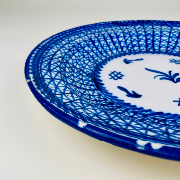 Manises ceramic wall art in the form of a large antique majolica plate, hand painted in cobalt blue on a white glazed body. The broad rim is filled with dense intersecting brush lines, contrasting with an open central medallion of botanical and geometric symbols. The signed Spanish charger has visible edge wear consistent with age.