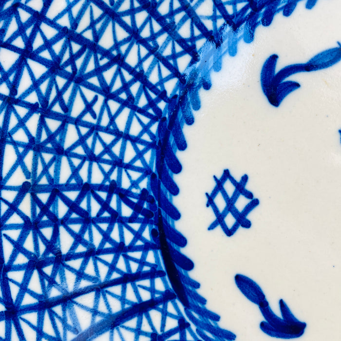 Antique Manises pottery charger made from tin glazed earthenware and painted in cobalt blue with an intricate geometric lattice. The broad border is built from overlapping diagonal, vertical, and radial lines, framed by darker blue bands around the rim and center. Small edge chips and glaze losses remain visible on this signed Spanish ceramic wall art.