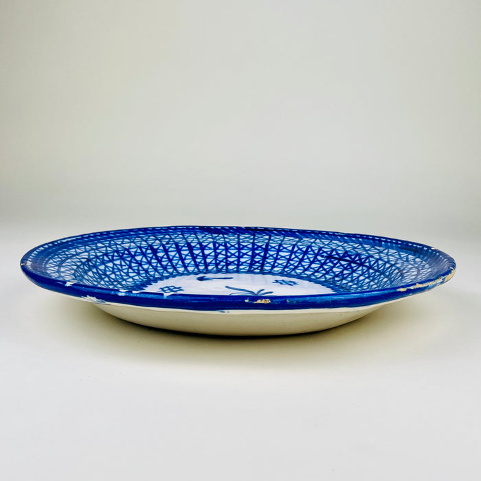 Signed Manises ceramic wall plate from early 20th century Spain, decorated by hand with saturated cobalt blue brushwork on a warm white glazed ground. Intersecting lines form a basket weave pattern around the rim, while the central medallion contains a plant sprig, leaf-like marks, and small crosshatched symbols suited to blue and white decor.