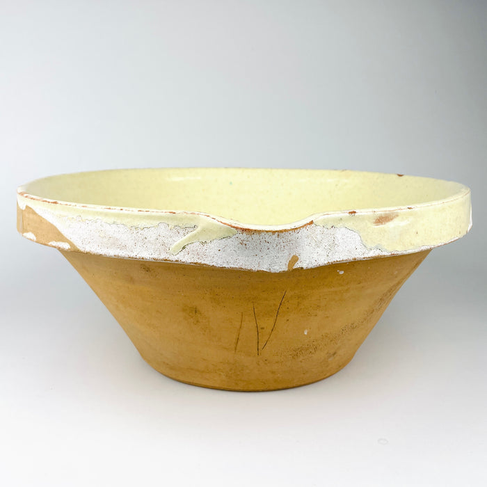 Early 20th century French terracotta tian bowl from Provence with a broad circular form, gently flared rim, small pouring notch, and pale yellow glaze over visible throwing rings. Glaze losses at the interior center and scattered chips around the rim reveal the natural clay body, creating authentic antique pottery character for rustic home decor.