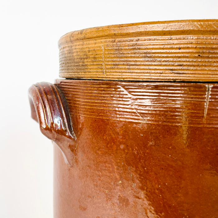 Antique French pottery storage vessel formed as a tall stoneware confit crock with a fitted lid and two compact side handles. Rich brown glaze covers the exterior and shifts from amber to chestnut, contrasting with the lighter grey-beige interior. The substantial jar can serve as a decorative vase, kitchen utensil holder, or grounded accent within French country decor and rustic home decor.