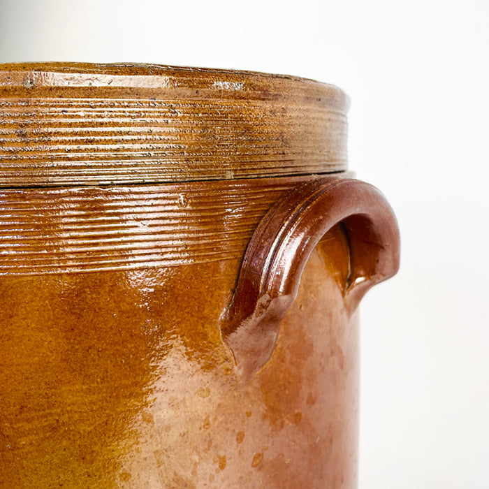 Early 20th-century French provincial confit pot in heavy brown glazed stoneware, complete with a fitted circular lid and two applied handles. Ribbed throwing lines around the lid and shoulder emphasise its handmade construction, while the pale interior glaze supports its original storage function. This antique crock brings aged pottery texture to farmhouse decor, Mediterranean decor, or cottage decor.