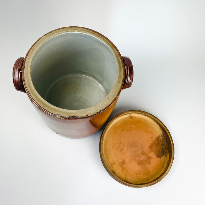 French country decor vessel formed as a large antique confit pot with its fitted lid and heavy stoneware construction. The warm chestnut-brown glaze, pale interior, compact ear handles, and ribbed wheel-thrown details create a restrained farmhouse character. Use this early 20th-century crock as a storage jar, utensil holder, dried flower vase, or sculptural centerpiece.