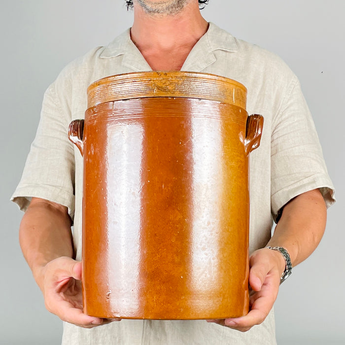 Large antique French confit pot in warm brown stoneware, with a fitted lid, robust cylindrical form, and paired ear handles. The glaze moves from dark chestnut near the shoulder to softer amber and clay tones lower on the body, with subtle wear from age and use. A substantial storage crock for vintage kitchen decor, French country decor, or styling with dried stems and branches.
