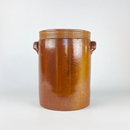 Antique French confit pot made from heavy wheel-thrown stoneware, retaining its matching lid and two applied side handles. The glossy chestnut-brown glaze varies naturally across the tall cylindrical body, while the ribbed neck and lid reveal the potter’s throwing process. A practical storage jar, utensil crock, or sculptural vessel for rustic home decor and vintage kitchen displays.