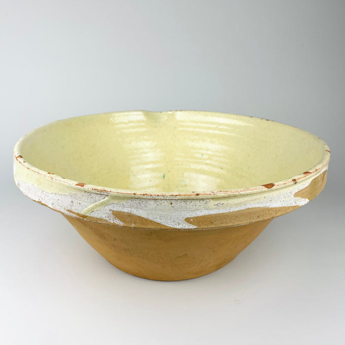 Large antique French tian bowl held from the side, showing the heavy conical profile, broad flared rim, unglazed ochre lower body, and pale glazed upper section. The substantial provincial terracotta form, pouring notch, and age-worn surface make this early 1900s Provence pottery especially effective as farmhouse decor or a sculptural centerpiece bowl.