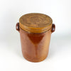 Large French stoneware confit pot dating to the early 20th century, with a tall storage crock form, matching fitted lid, and twin pierced handles. Brown, amber, and russet glaze tones cover the exterior, while a pale grey-beige glaze lines the inside. The robust vessel suits antique and vintage collections, farmhouse decor, cottage decor, or a decorative dried botanical arrangement.