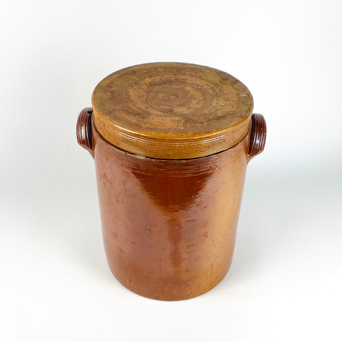 Large French stoneware confit pot dating to the early 20th century, with a tall storage crock form, matching fitted lid, and twin pierced handles. Brown, amber, and russet glaze tones cover the exterior, while a pale grey-beige glaze lines the inside. The robust vessel suits antique and vintage collections, farmhouse decor, cottage decor, or a decorative dried botanical arrangement.