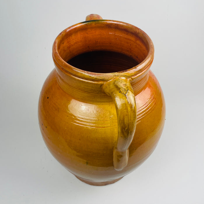 Antique Spanish earthenware storage jar with two handles, workshop glazed in a warm amber tone with a distinct glaze break and drip effects. The lower terracotta body shows age-related scuffs and marks, while the upper glazed section reflects light strongly, making it a statement ceramic vessel for console decor or entryway styling.