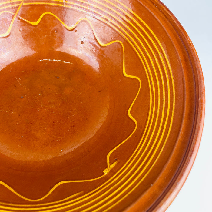 The broad circular form of the antique French tian bowl is defined by an amber brown glazed interior, a thick everted rim, repeated yellow bands, and a freehand undulating border. This substantial early 20th century terracotta bowl can anchor a dining table as a decorative centerpiece or hold fruit in a French country kitchen display.