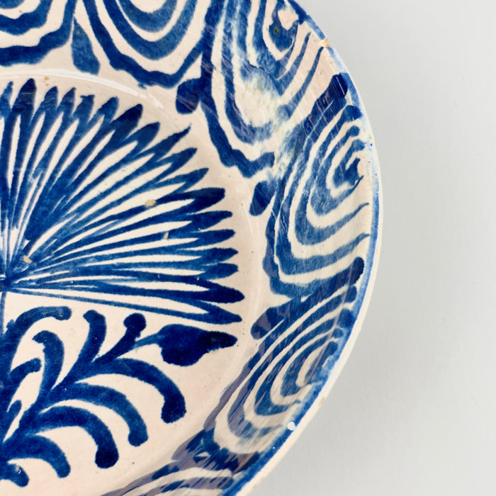 Close-up of rim section showing cobalt spiral wave motif and visible rim chip, illustrating the natural wear and character of this antique Spanish pottery while emphasizing the hand-painted detailing and glaze variation.