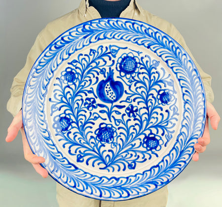 Spanish Fajalauza style platter with a hand painted pomegranate at the center, surrounded by cobalt blue flowers, curling stems, and a dense vegetal border on a white glazed ground. This contemporary ceramic platter can be displayed as a wall hanging plate or used as a large serving charger in Mediterranean decor, Spanish decor, or rustic dining interiors.