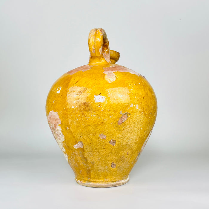 Reverse view of a large antique French cruche pot from Provence, showing the strongly curved earthenware body and sculptural overhead handle. Honey-yellow glaze, extensive areas of rose terracotta and pale mineral deposits create a richly weathered pottery surface for collected Mediterranean decor.