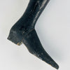 Antique French mannequin leg formed from heavy carved wood, with a long boot joined around the ankle and a shaped heel supporting the object upright. A visible gap follows the historic construction seam, accompanied by scattered small holes and broad rubbing to the black finish. The timeworn surface adds texture to bookshelf decor.