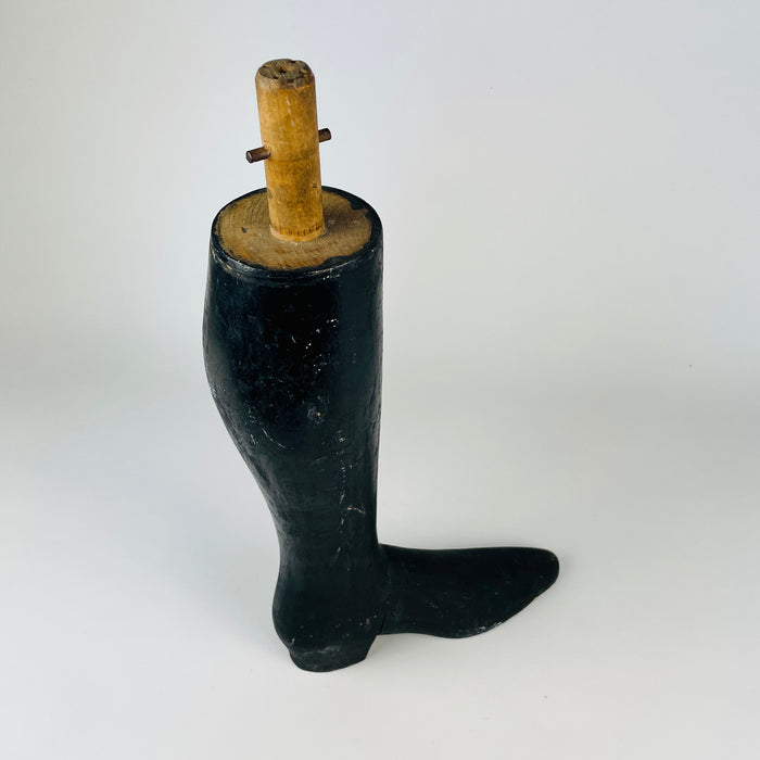 Antique French wooden mannequin leg dating to circa 1870–1890, carved with a slender calf, sharply defined ankle and unusually long black boot. Heavy wear exposes pale wood through the dark painted finish, while the tall silhouette and surviving mounting hardware retain the character of a working trade object for dark academia decor.