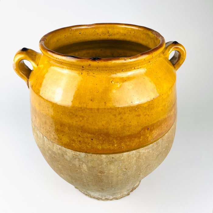 Antique South West French confit pot formed in terracotta with paired applied handles, a rounded shoulder and a narrow foot. The mustard yellow glaze is concentrated around the mouth and upper body, contrasting with the pale, worn lower surface and suiting collected farmhouse decor.