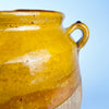Late 19th century French terracotta confit pot with an applied ear handle rising from the shoulder below a rolled rim. The upper section carries luminous yellow and honey glaze, while the lower body remains weathered and matte, creating an organic modern decor statement with historic function.