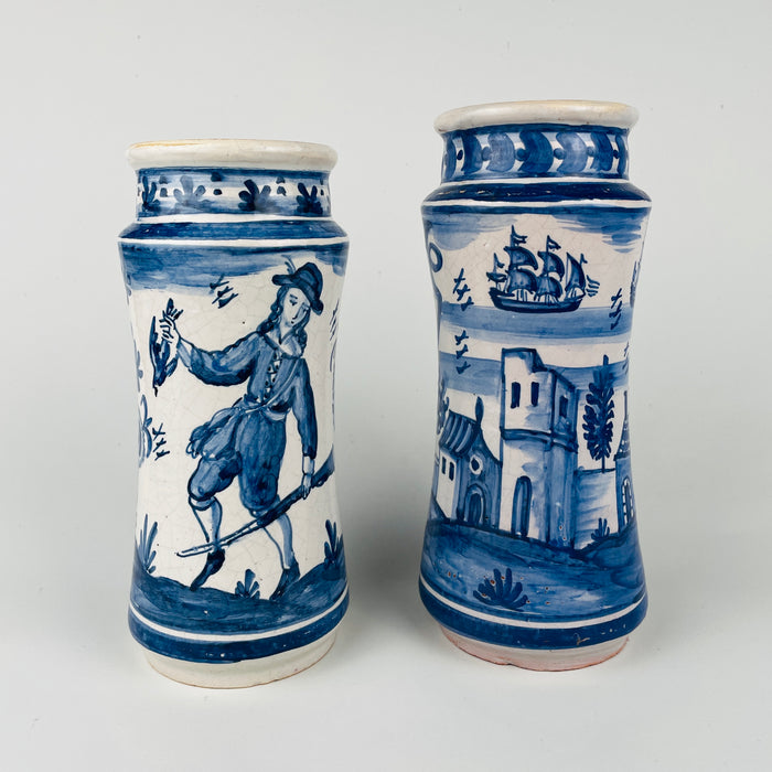 Set of two antique Spanish faience apothecary jars (albarellos) in white tin glazed earthenware with cobalt blue hand painted labels and scenic motifs. One jar reads “VALLERIANA” and shows a Spanish galleon sailing ship with coastal architecture; the other reads “EUCALIPTO” with a standing figure and floral border. Mediterranean decor storage for herbs, utensils, or dried flowers.