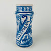 Antique Spanish blue and white faience pharmacy jar labeled “VALLERIANA,” decorated with a hand painted Spanish galleon sailing ship, buildings, and landscape details in cobalt blue on white tin glazed ceramic. Cylindrical albarello form with flared rim and unglazed foot ring, showing authentic glaze crazing and kiln wear. Coastal decor and nautical decor accent for shelves, consoles, and kitchen storage.