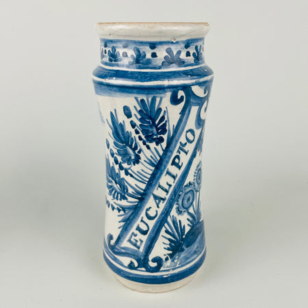 Antique Spanish blue and white faience apothecary jar labeled “EUCALIPTO,” featuring a hand painted standing figure with foliate scrollwork and border bands in cobalt blue over a white tin glazed ground. Traditional albarello form used for pharmacy botamen, suitable today as a Mediterranean decor vessel for countertop utensils or dried botanicals. Surface shows glaze crazing and small edge wear consistent with age.
