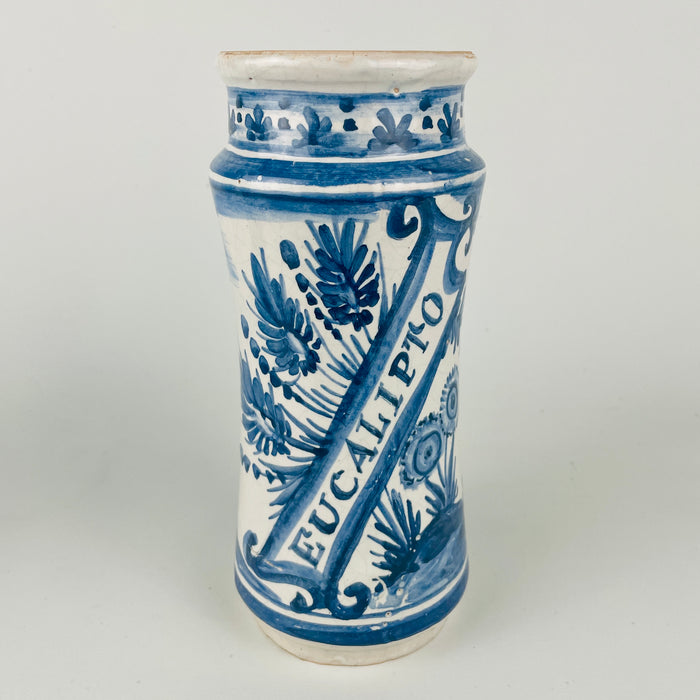 Antique Spanish blue and white faience apothecary jar labeled “EUCALIPTO,” featuring a hand painted standing figure with foliate scrollwork and border bands in cobalt blue over a white tin glazed ground. Traditional albarello form used for pharmacy botamen, suitable today as a Mediterranean decor vessel for countertop utensils or dried botanicals. Surface shows glaze crazing and small edge wear consistent with age.