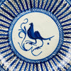 Detail image of central blue bird painted in stylized brush strokes, encircled by white ring and surrounded by radiating blue decorative lines on antique ceramic plate.