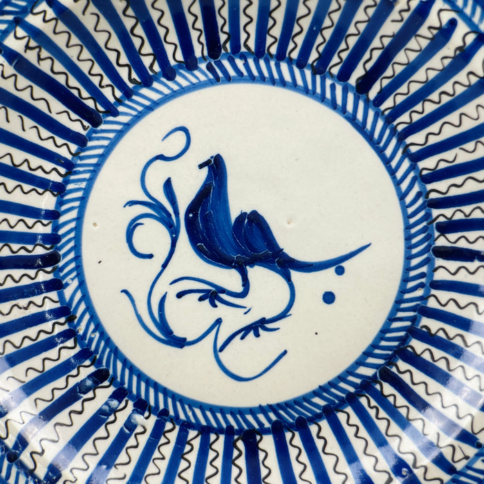 Detail image of central blue bird painted in stylized brush strokes, encircled by white ring and surrounded by radiating blue decorative lines on antique ceramic plate.