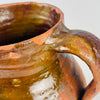 Antique Castilian pottery jar with one of its tall hand applied loop handles visible against the rounded terracotta body. Honey brown glaze covers the shoulder and upper section while the warm red clay remains exposed below, creating the worn two tone character associated with Spanish vernacular pottery and Mediterranean decor.