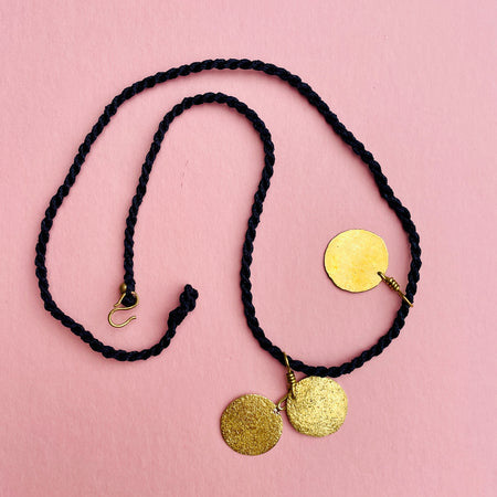 Front view of the gold-tone disc necklace from the Aurelia Collection by ROC Objects, showing three hammered brass pendants on a braided black cord. The necklace measures 49.5 cm and sits as a minimalist layering piece, perfect for boho and contemporary styling.

Angled close-up highlighting the ha