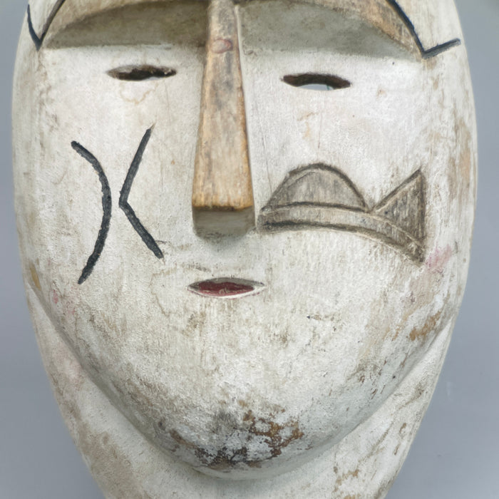 Face-forward image of African ceremonial mask with sculpted nose ridge, black cheek incisions, and white pigment finish, inspired by Fang and Kwele art, shown as a decorative tribal wall sculpture or folk decor.

