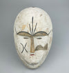 Hand-carved African ceremonial mask featuring angular facial lines, whitewashed surface, and narrow eyes, reflecting stylistic elements of Fang or Kwele traditions, photographed for display as tribal wall art or global folk dec