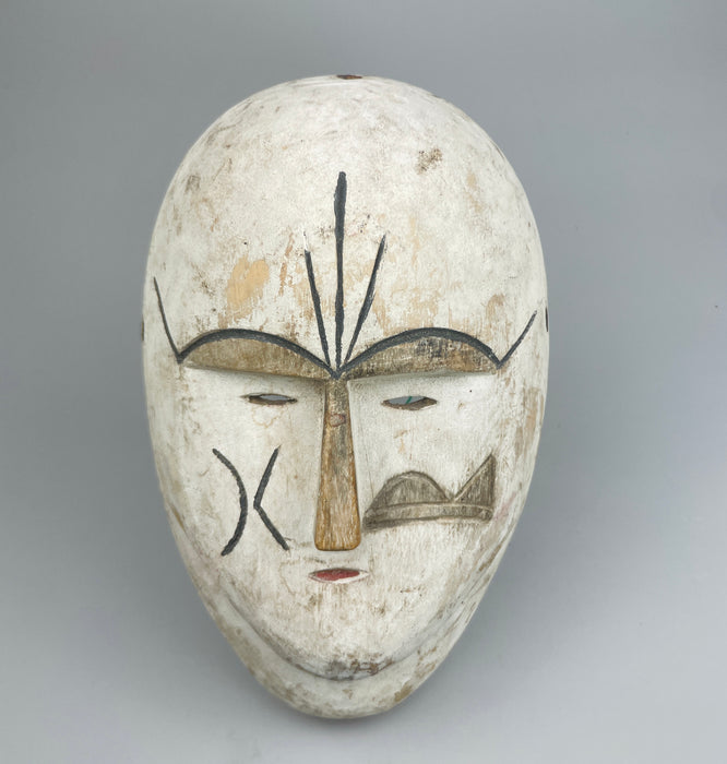 Hand-carved African ceremonial mask featuring angular facial lines, whitewashed surface, and narrow eyes, reflecting stylistic elements of Fang or Kwele traditions, photographed for display as tribal wall art or global folk dec