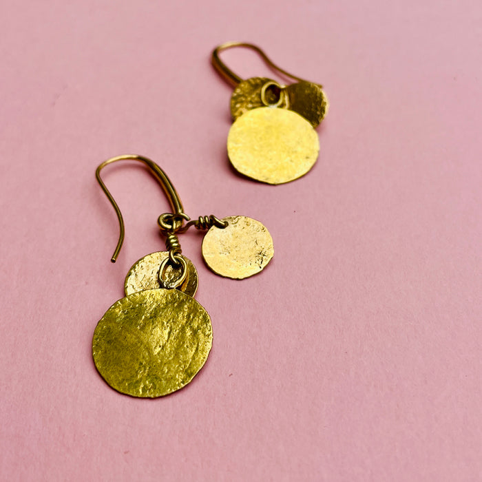 Pair of hammered coin drop earrings shown with reflective light on textured surfaces. Gold-tone alloy discs shimmer against nickel-free hooks, highlighting their versatile styling for resort fashion, capsule jewelry collections, or everyday lightweight wear with Mediterranean design influence.