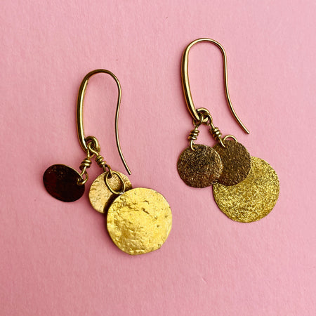 Close-up of artisan-made hammered coin dangle earrings in gold-tone alloy, suspended from nickel-free hooks. The discs, measuring 1.9 cm and 0.6 cm, reflect light with a warm golden glow, offering a handcrafted finish perfect for boho-chic styling, casual everyday wear, or layering with other gold jewelry.