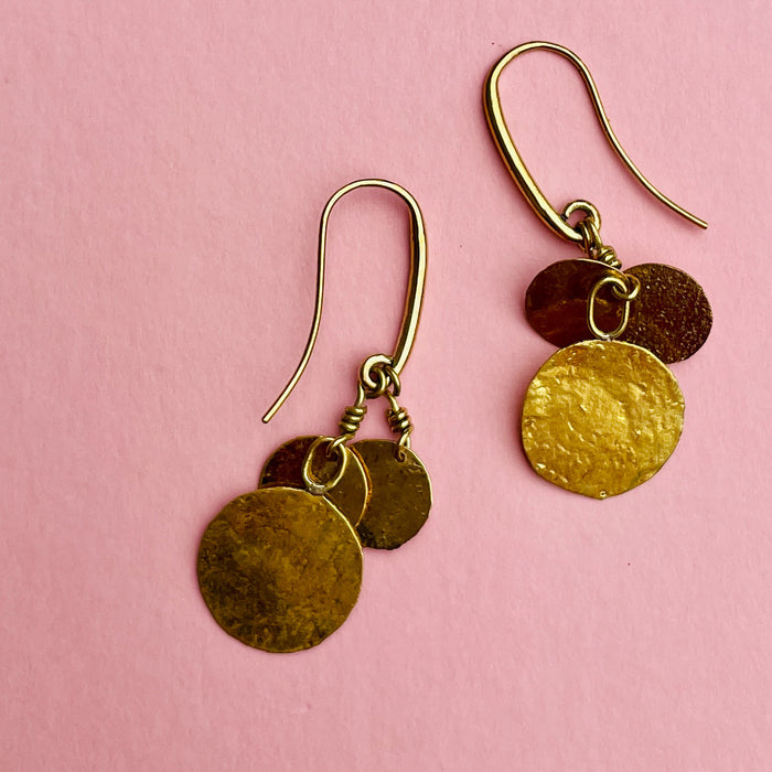 Overhead display of hammered coin earrings, featuring three golden discs per earring on nickel-free hook wires. Their reflective hammered finish creates fluid shimmer, making them versatile jewelry accents suitable for everyday wear, festivals, or modern Mediterranean fashion collections.