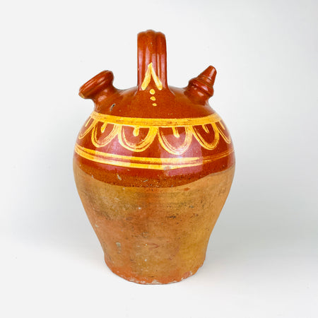 Antique terracotta water jug from northern Spain, dating to the early 20th century, made in the traditional botijo form with a rounded body, arched handle, wide filling mouth, and narrow drinking spout. The upper half is glazed in a warm chestnut brown and hand painted with yellow scalloped decoration, while the lower body remains unglazed red clay. This Spanish folk pottery vessel can be styled in Mediterranean decor, farmhouse decor, and vintage home decor.