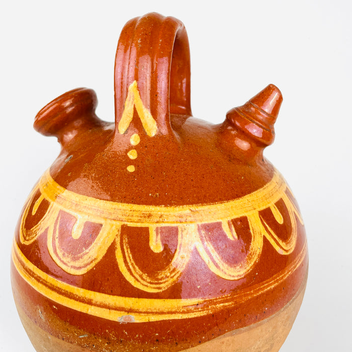 Northern Spanish botijo in red terracotta with partial brown glaze and hand painted yellow banding, made as a traditional water jug for cooling and storing water. The broad shoulder, tall loop handle, open mouth, and tapered spout define the classic functional form, while the unglazed lower section preserves the porous clay character associated with antique Spanish pottery. This decorative vessel suits rustic decor, wabi sabi decor, and country home pottery displays.