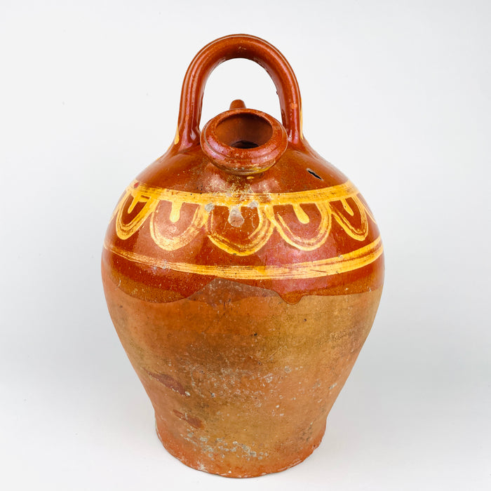 Antique Spanish folk pottery vessel in the form of a botijo, made in terracotta and decorated with hand painted yellow lines and repeated curved motifs over a glazed brown ground. The lower half is left in natural clay, creating contrast between glossy glaze and matte porous earthenware. Measuring approximately 33 cm high, this water jug has an estimated capacity of about 3 L and can be displayed as a decorative terracotta vessel in farmhouse decor, Mediterranean decor, or vintage home decor.