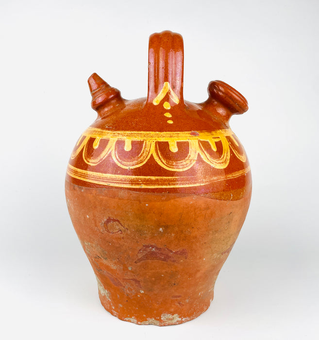 Northern Spain antique botijo in red clay, early 20th century, with a rounded shoulder, tapered body, and attached vertical handle spanning the top of the vessel. The upper body is glazed and painted in a yellow on brown palette, while the lower body remains unglazed and visibly worn, showing the honest surface of traditional utility pottery. This Spanish terracotta water jug is well suited to rustic decor, country pottery displays.