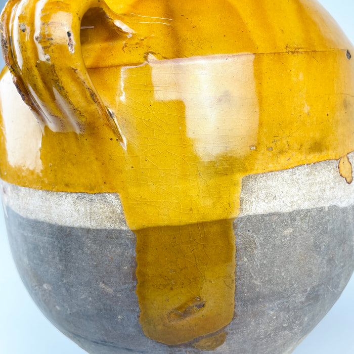 Detail of the antique French confit pot showing an applied terracotta loop handle, glossy ochre yellow glaze and a broad vertical glaze run extending onto the unglazed lower body. Fine crazing, worn edges and small exposed clay areas are clearly visible, emphasizing the hand-finished surface and genuine age of this 19th century French pottery vessel.