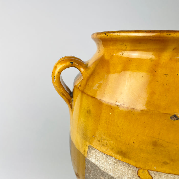 Applied loop handle and shoulder detail on an antique French terracotta confit pot, with mustard yellow glaze following the hand-shaped curves of the handle and neck. Small glaze chips, abrasions and exposed clay along the handle edges reveal authentic wear, while the rounded shoulder and flared rim retain the distinctive silhouette associated with traditional French pottery jars.