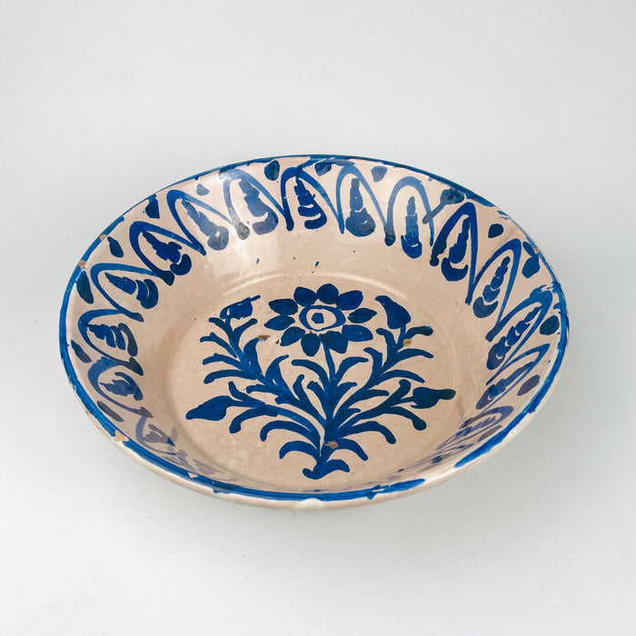 Spanish Granada lebrillo bowl in antique Fajalauza earthenware, decorated with a stylized blue flower and branching foliage in the center, surrounded by a rhythmic blue border painted freehand around the interior wall. The cream-white glaze shows light crazing and age wear, with small chips along the rim. This antique Spanish pottery bowl fits Mediterranean decor, farmhouse decor, and decorative wall bowl collections focused on regional ceramic traditions.