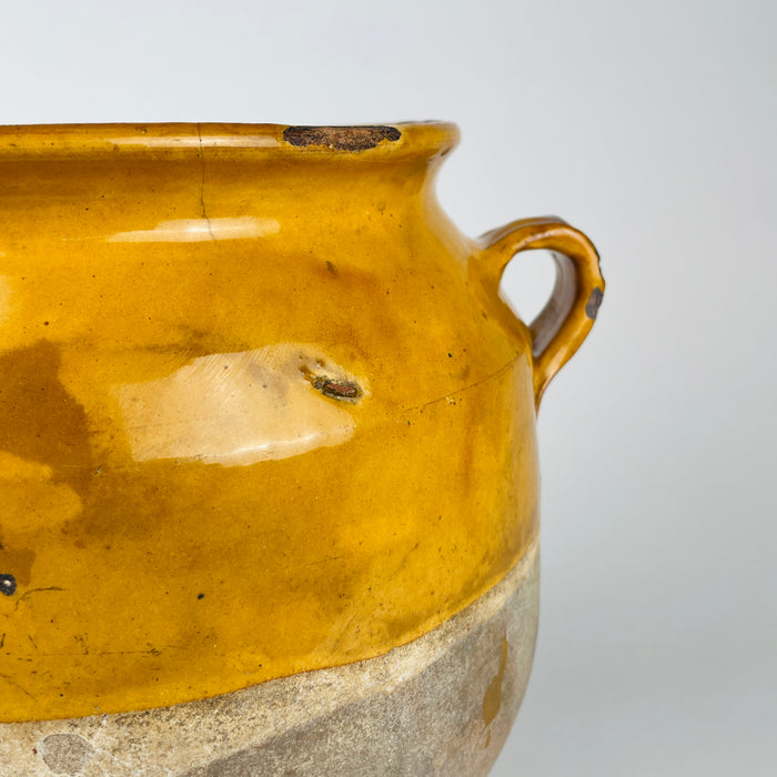Surface detail of the antique French confit pot showing the saturated honey yellow glaze, subtle speckling, age crazing and a fine horizontal linear fissure across the upper body. The transition from glazed shoulder to raw lower terracotta is visible below, together with small glaze losses that contribute to the timeworn surface of this 19th century French vessel.