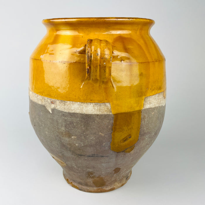 French antique confit pot with a broad rounded body, narrow tapered foot and paired intact handles, made from terracotta and partially covered in honey yellow glaze. The exposed lower body retains a heavily aged matte surface, while the glossy upper glaze shows small chips and wear from long use, giving this 19th century pottery jar strong rustic home decor character.