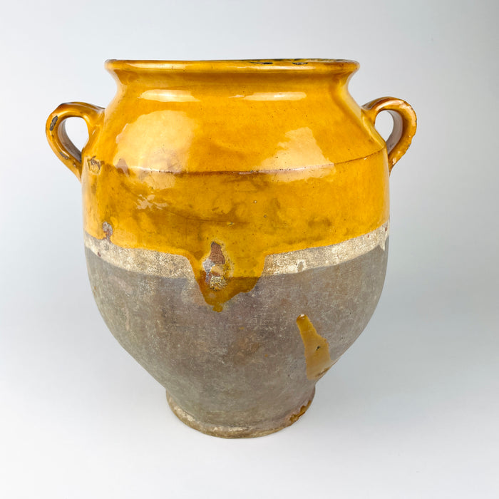 Antique French terracotta confit pot from the 19th century, with a lustrous yellow ochre glazed upper section set against the unglazed pale grey-beige lower body. Twin loop handles, a flared lip and a pronounced taper toward the foot give the vessel a sculptural silhouette, while chips, scratches and glaze variation preserve its aged French country decor appeal.