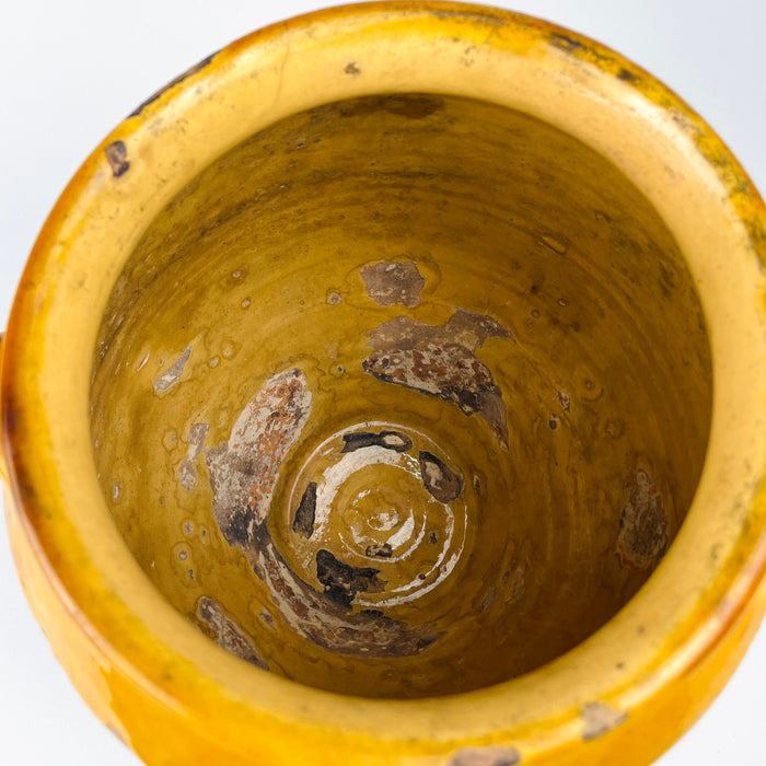 Antique French confit pot with its strongly tapered lower body and small circular foot clearly visible beneath the wide rounded shoulder. The lower terracotta is intentionally unglazed and heavily abraded to a pale grey-beige surface, contrasting with the yellow glazed upper section and emphasizing the sculptural shape of this substantial 19th century pottery jar.