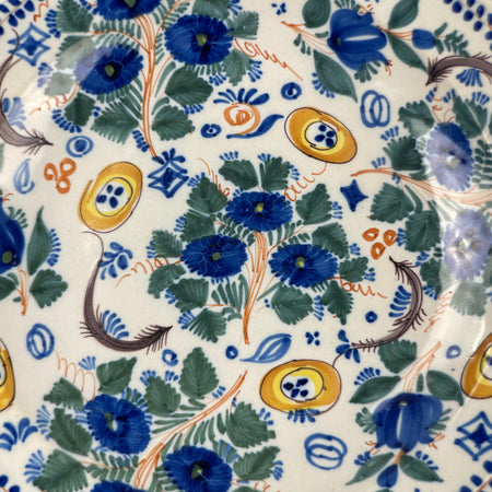 Close-up of blue flower and green foliage design on antique tin-glazed Spanish plate, highlighting painterly detail, crisp brushwork, and vibrant color against the white ground for rustic or global décor.