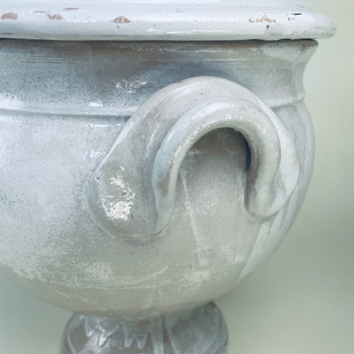 Large white ceramic tureen from France, designed with an urn shaped body, broad pedestal foot, two curved handles, and a high domed cover. The worn white glaze and exposed terracotta around the lower edge give this decorative bowl a layered surface for French country decor, cottage decor, or neutral vintage home decor.